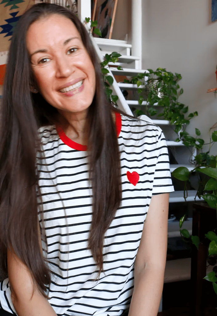 Cara Carmina sonriente, con cabello largo y lacio, vestida con camiseta de rayas blancas y negras con un corazón rojo, rodeada de plantas en un ambiente luminoso.